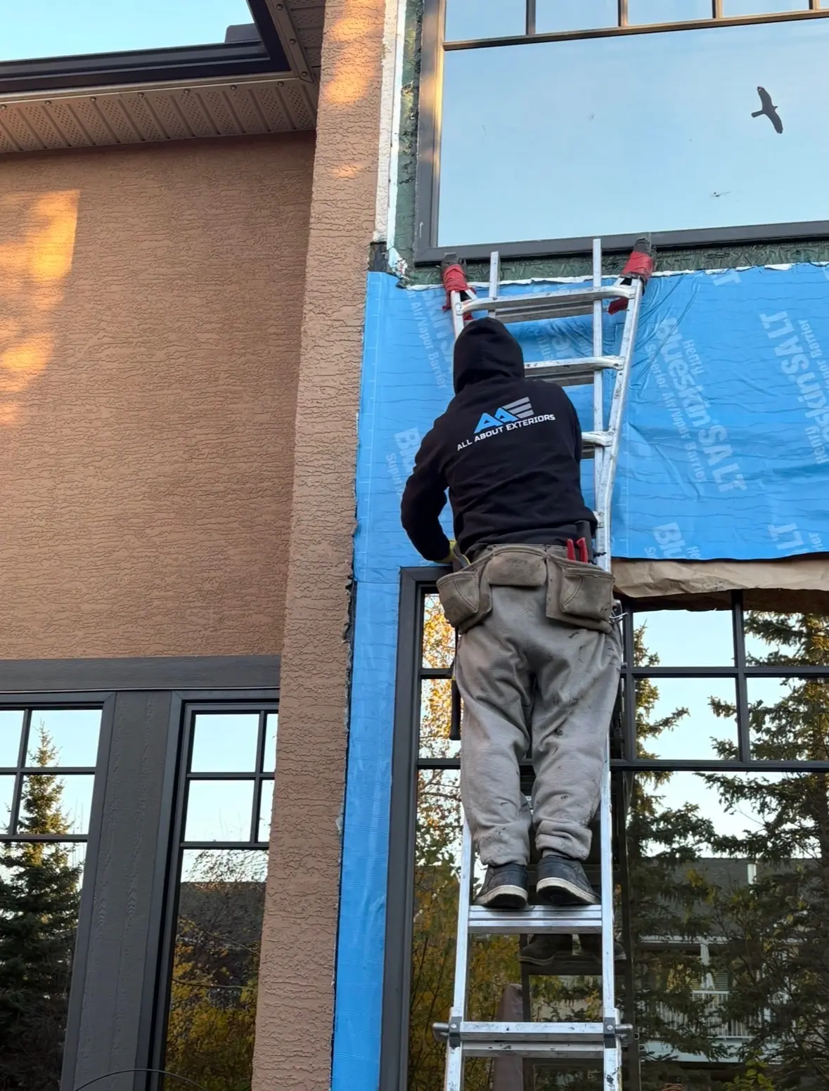 All About Exteriors crew member on a ladder installing Henry Blueskin air and vapor barrier on a stucco home