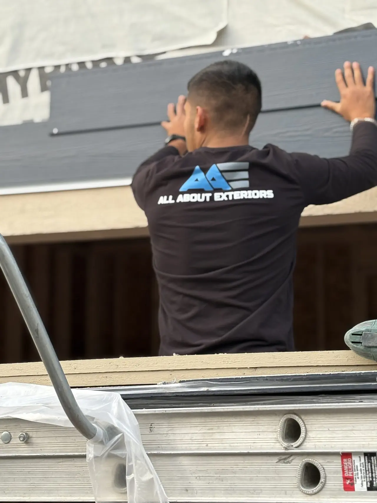 All About Exteriors installer fitting a James Hardie siding panel from a ladder jack