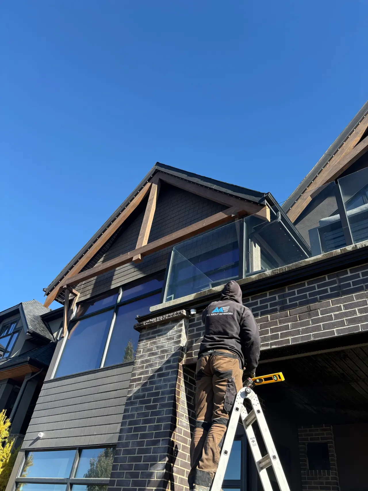 All About Exteriors crew member on a ladder below a timber gable on a dark brick and shingle-clad home
