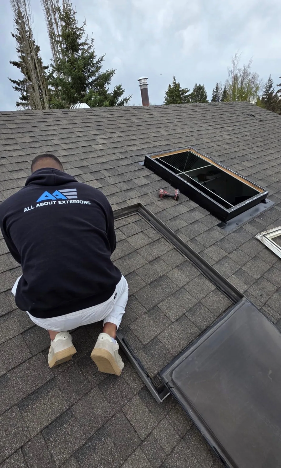 All About Exteriors roofer kneeling on a shingle roof beside an opened skylight frame