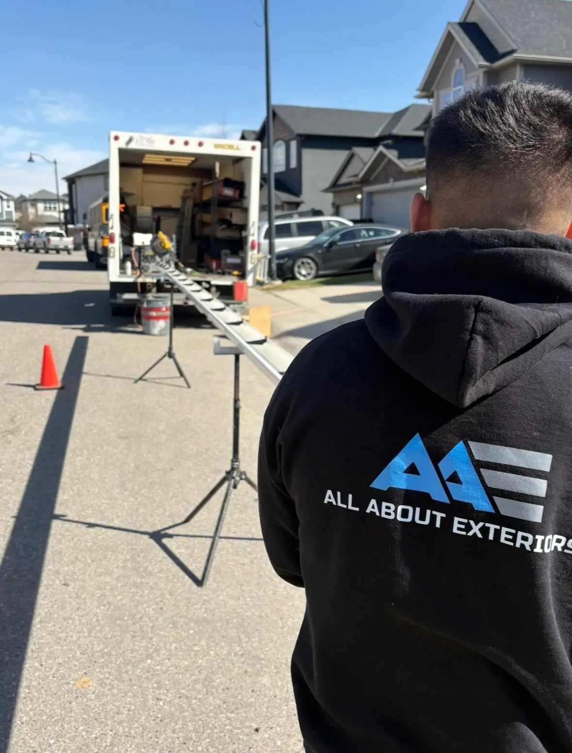 All About Exteriors crew member in a company hoodie looking down a long gutter run set on stands beside the open box truck on a residential street
